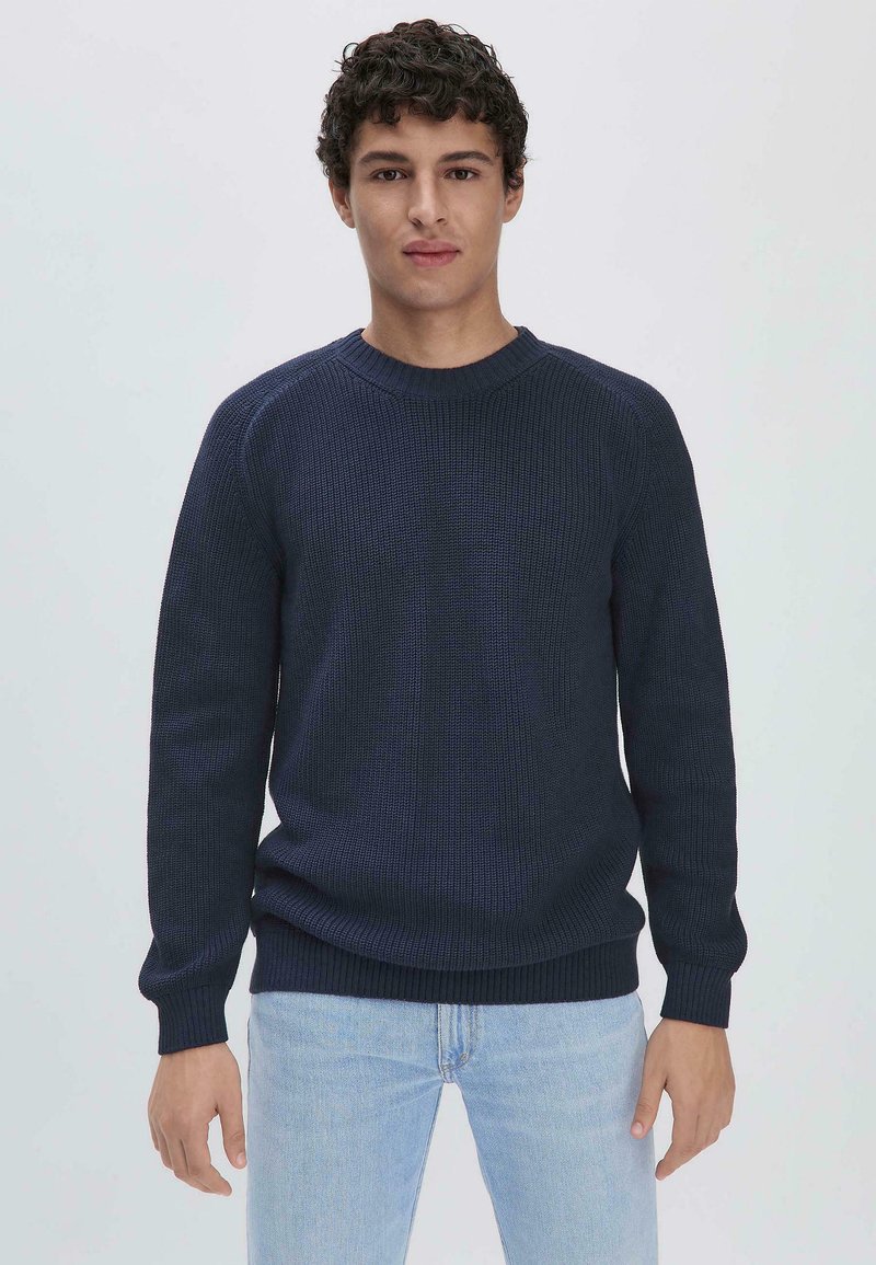 Marineblauer gerippter Strickpullover mit Rundhalsausschnitt, langen Ärmeln und strukturiertem Stoff. Kombiniert mit hellblauen Jeans, präsentiert vor einem einfarbigen Hintergrund.