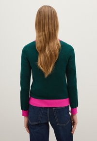 Maglione di lana verde con polsini e orlo rosa, caratterizzato da un design semplice e aderente con una texture a coste. Indossato con jeans in denim blu.