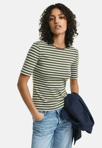 Stribet kortærmet top i olivengrøn og hvid, med en rund hals og en tætsiddende design, parret med lyseblå jeans og en marineblå stribet jakke.