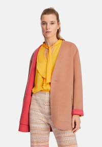 Strickmantel in Hellrosa mit roten Akzenten, über einer gelben Bluse mit Rüschen getragen. Gemusterte, taillierte Hose mit geometrischem Design.