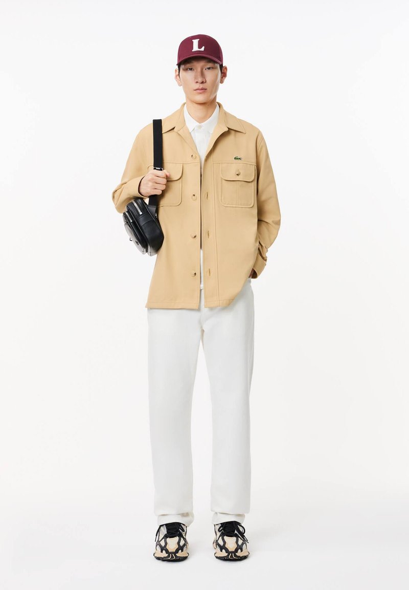 Beige Knopfjacke mit zwei Fronttaschen, weißem Hemd, weißen Hosen, schwarzer Tasche und gemusterten Sneakern mit schwarzen und beige Akzenten. Kastanienfarbene Mütze.
