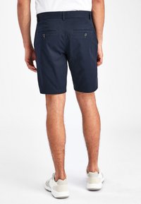 Pantaloni scurți bleumarin cu o textură netedă, până la mijlocul coapsei, două buzunare pe spate cu nasturi și un mânere pentru curea la talie. Se văd adidași.