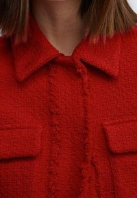 Red textured jacket featuring a collared neck, frayed accents down the front, and patch pockets on either side.