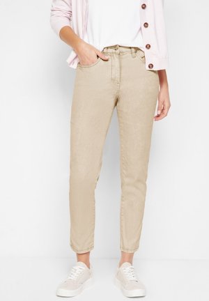 Jean beige taille haute avec une jambe fuselée, présentant des poches avant, une fermeture à bouton et une texture douce et lisse, assortis à des chaussures blanches.