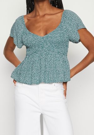 Mujer con blusa verde floral de manga corta y peplum y pantalones blancos de tiro alto, con las manos en las caderas, de pie frente a un fondo liso.