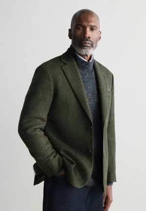 Blazer de lana verde oscuro con un acabado texturizado, diseño de un solo botón y botones marrones, superpuesto a un suéter de cuello alto gris.