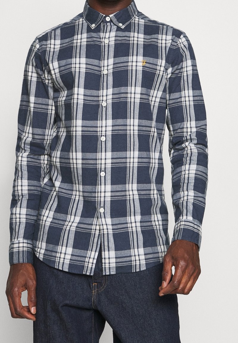 Homme portant une chemise à carreaux bleu marine et blanc à manches longues et un jean bleu foncé.