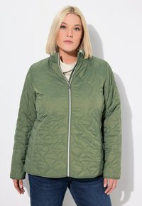 Giacca trapuntata verde con colletto alto e chiusura con zip. Il tessuto presenta un motivo esagonale testurizzato. Indossata sopra una maglia chiara.