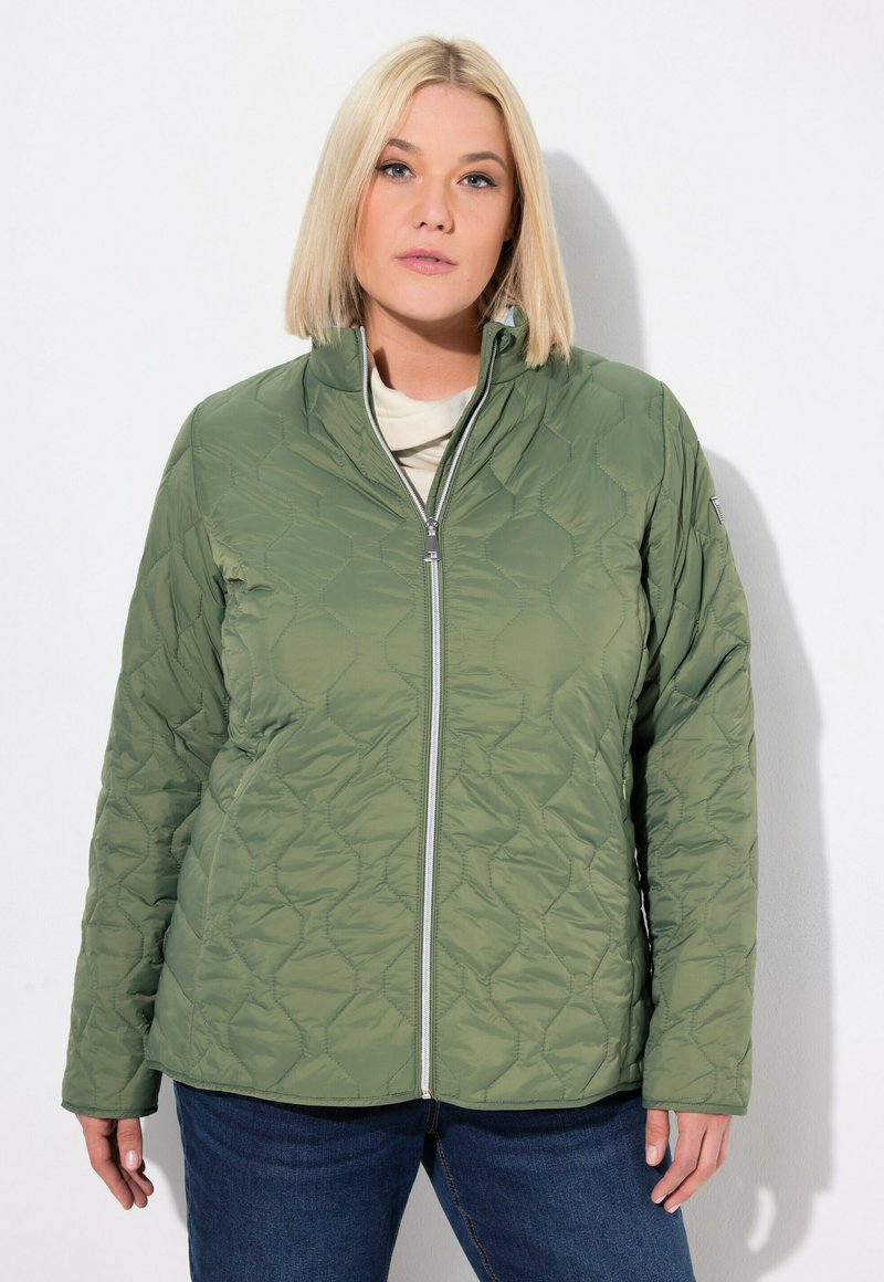 Giacca trapuntata verde con colletto alto e chiusura con zip. Il tessuto presenta un motivo esagonale testurizzato. Indossata sopra una maglia chiara.