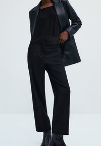 Zwart leren blazer over een zwarte top, gecombineerd met hoge zwarte broek. Heeft een soepele textuur en een getailleerde pasvorm. Zwarte enkellaarzen.