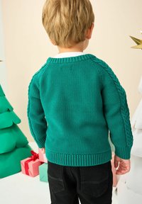 Kind mit kurzen blonden Haaren, das einen türkisfarbenen Strickpullover und schwarze Hosen trägt, steht mit dem Rücken zur Kamera in der Nähe von eingewickelten Geschenken und Papierbäumen.