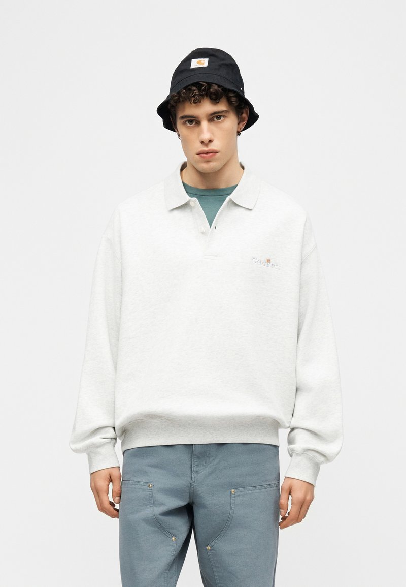 Junger Mann trägt einen weißen Langarm-Pullover, blaue Hosen mit Fronttaschen, ein türkisfarbenes Hemd darunter und einen schwarzen Bucket-Hut mit Logo.