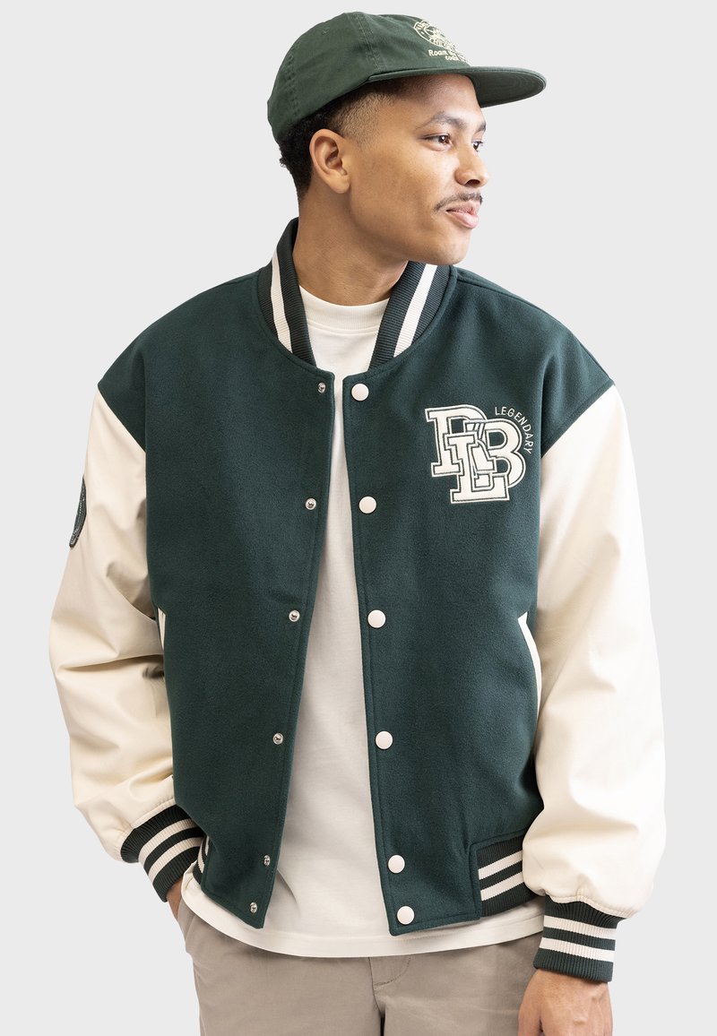Green and cream varsity jacket with snap buttons, ribbed cuffs, and a logo on the chest. Plain white shirt worn underneath.