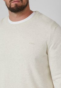 Ein bärtiger Mann trägt einen hellbeigen Rundhalspullover über einem weißen T-Shirt, mit einem dezent gestickten Logo auf der Brust.