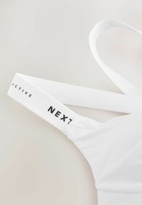 Brassière de sport blanche avec sangle élastique portant l'inscription noire « NEXT ACTIVE » sur un fond clair uni.
