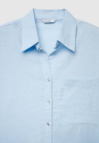 Chemise bleu clair à boutons avec col pointu et poche poitrine, montrant les trois premiers boutons et une étiquette portant la mention "CALLIOPE S" à l'intérieur du col.