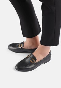 Zwarte leren instappers met gouden ronde accenten, platte zolen en een gladde textuur. Gecombineerd met op maat gemaakte zwarte broeken, wat een casual ontwerp benadrukt.