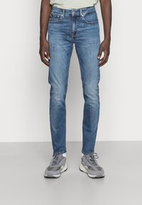 Jeans de mezclilla azul claro con corte slim, que presentan un clásico diseño de cinco bolsillos y un ligero desgastado. Combinados con zapatillas deportivas grises.