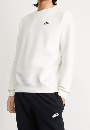 Osoba w białej bluzie Nike i czarnych spodniach Nike, stojąca z jedną ręką w kieszeni na jednolitym tle.