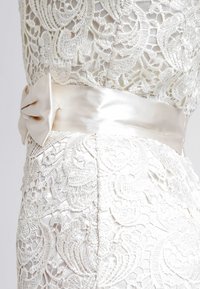 Spetsklänning med intrikata blommönster i elfenben, med ett brett satininspirerat bälte knutet i en rosett i midjan som kompletterar det strukturerade tyget.