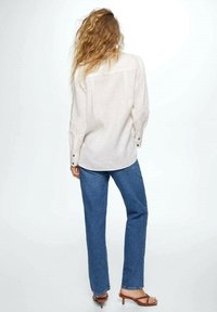 Kvinde med bølget hår iført en løs hvid langærmet skjorte, blå lige ben jeans og brune hælede sandaler, vendt væk.