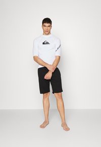 White rash guard with black logo, 3/4 sleeves; paired with black shorts. Model stands barefoot, arms crossed, facing forward.