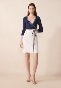 Haut en wrap marine avec des bordures blanches et un décolleté plongeant, accompagné d'une jupe plissée blanche, mise en valeur par un lien à la taille. Des sandales transparentes complètent le look.