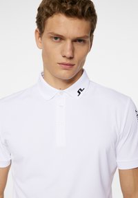 Valkoinen polo-paita klassisella kauluksella, jossa on kolme nappia ja pieni musta logo kauluksessa. Liukas, kevyt kangas. Lyhyet hihat.