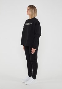 Felpa nera oversize con un logo grafico sul petto, abbinata a pantaloni neri a gamba affusolata e sneakers bianche. Tessuto dalla consistenza liscia.
