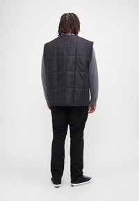 Melna izolēta veste ar kvadrātveida quilted rakstu, bez piedurknēm, ar augstu apkakli, uzvilkta virs pelēka garām piedurknēm krekla un melniem biksēm.