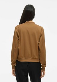 Femme aux longs cheveux noirs portant une veste bomber marron et un pantalon noir, de dos contre un fond clair et uni.