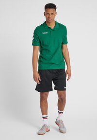 Grøn polo t-shirt med krave, korte ærmer og hvidt logo; parret med sorte shorts. Model iført grå sko med røde detaljer.