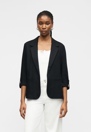 Kvinde med flettet hår iført guld ringearreringe, en sort blazer med oppullede ærmer, hvid top og hvide bukser mod en ensfarvet baggrund.