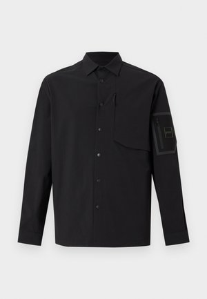 Chemise noire à manches longues avec fermeture par boutons sur le devant, rabat de poche poitrine zippé, et poche zippée avec patch logo sur la manche gauche.