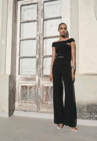 Schwarzer, schulterfreier Jumpsuit mit weitem Bein, figurbetontem Oberteil und silbernem Gürtel. Modell trägt weiße Flip-Flops und steht neben Holztüren.