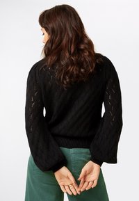 Suéter negro de punto con un patrón calado y mangas voluminosas, combinado con pantalones de pana verdes. Dobladillo y puños acanalados.