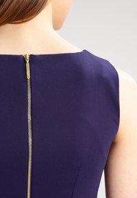 Robe marine avec un décolleté simple et une fermeture éclair dorée dans le dos, mettant en valeur un tissu lisse et structuré avec peu de détails.