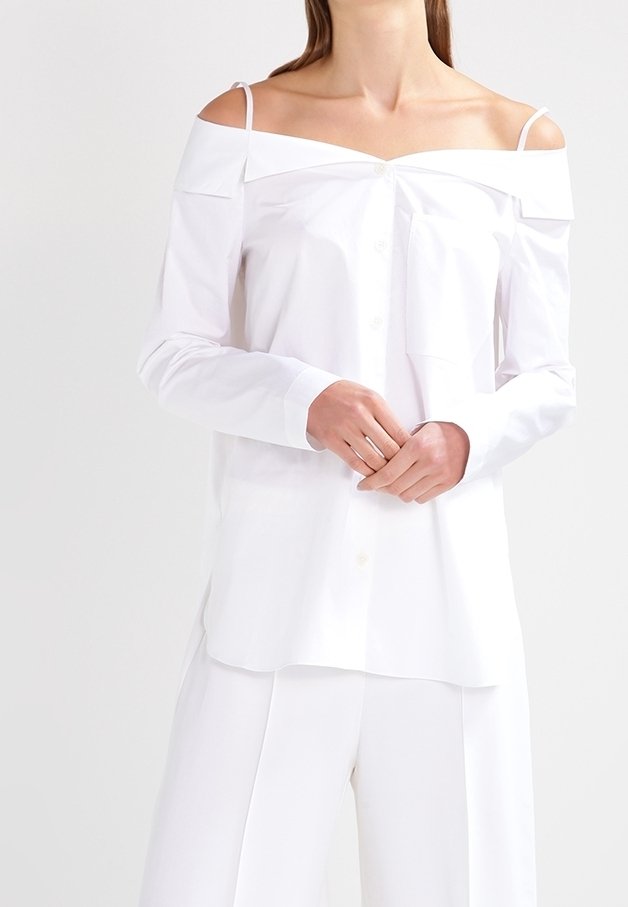 Model iført en off-shoulder hvid skjorte med lange ærmer, knaplukning foran og et slankt, minimalistisk design mod en lys baggrund.