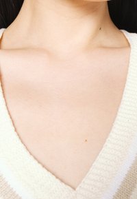 V-paita, jossa on teksturoitu, kermaisen värinen neulos. Kaula-aukoissa näkyy sileä iho, jossa on pieni luomi solisluun alueella.