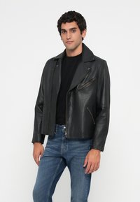 Chaqueta de motorista de cuero negra con un cuello ancho, cremallera frontal asimétrica y bolsillos con cremallera. Se lleva con una camisa negra y jeans azules.
