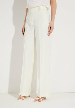 Frau trägt hoch taillierte, weit geschnittene weiße Hosen mit Gürtelschlaufen und weiße offene Sandalen mit Absatz, steht vor weißem Hintergrund.