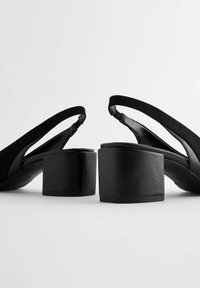 Musta kiilakorkoiset sandaalit, joissa on paksu lohkokorko, mokkanahkainen päällinen ja avonainen kantapääremmi. Silkkiset nahka-yksityiskohdat korostavat kengän modernia muotoilua.