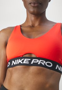 Persona atlética vestida con un sujetador deportivo rojo brillante con banda negra de Nike Pro, mostrando hombros tonificados y parte superior del torso contra un fondo liso.