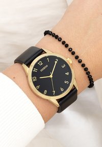 Montre dorée avec un cadran noir, de grands chiffres blancs et un bracelet en cuir noir. Accompagnée d'un bracelet en perles noires.