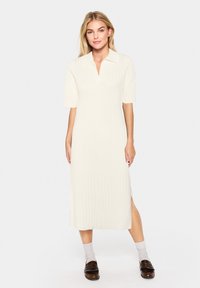 Robe midi de couleur crème claire avec texture côtelée, col et manches courtes ; présente une fente latérale et une silhouette droite. Portée avec des mocassins sombres.