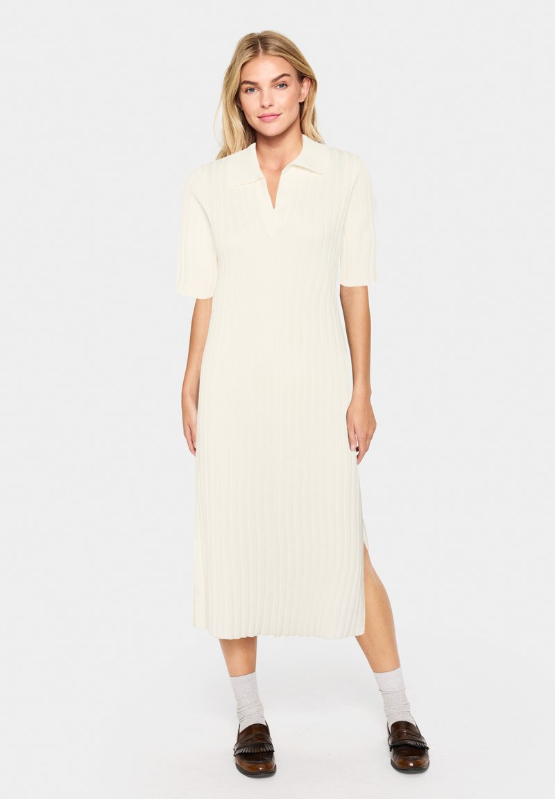Robe midi de couleur crème claire avec texture côtelée, col et manches courtes ; présente une fente latérale et une silhouette droite. Portée avec des mocassins sombres.