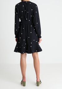 Robe noire à pois blancs, manches longues, taille cintrée. Jupe évasée avec un ourlet au-dessus du genou, tissu texturé. Chaussures à talons verts.