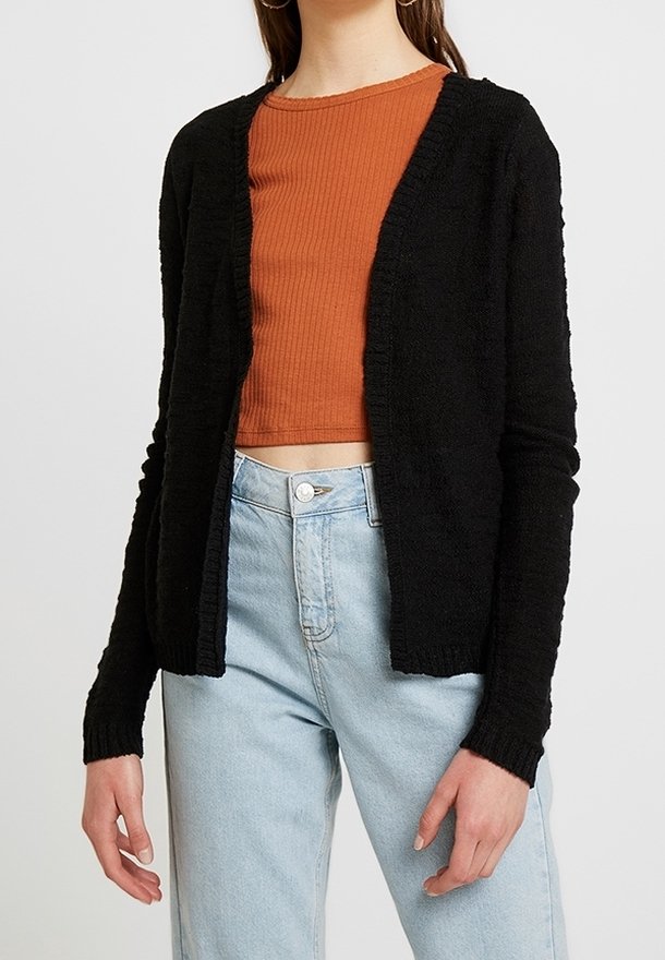 Black knitted cardigan with open front, long sleeves, and ribbed texture, worn over a rust-colored crop top and light blue high-waisted jeans.