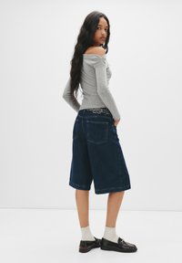 Graues, schulterfreies Langarmoberteil kombiniert mit dunkelblauen Jeansculottes. Ein silberner Nieten-Gürtel, graue Socken und schwarze Loafers runden den Look ab.