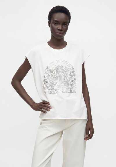 Femme portant un haut blanc sans manches avec un motif en strass et le texte "FOR REALITY I WANT MAGIC," associé à un pantalon blanc sur un fond uni.